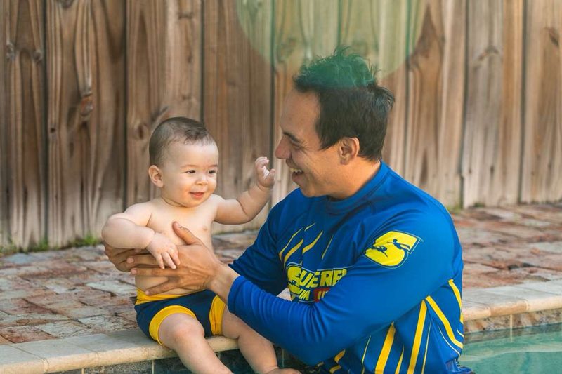 Coach in a Superhero Swim shirt holding a toddler at the poolside